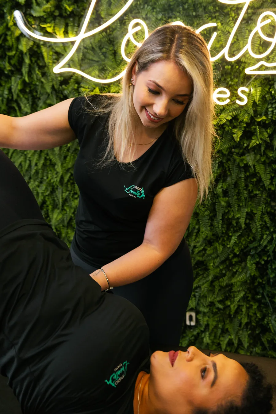 Pricila Lovato ajoelhada no chão do studio, sorrindo enquanto orienta uma aluna deitada no tapete, com o neon do Studio Lovato Pilates ao fundo.