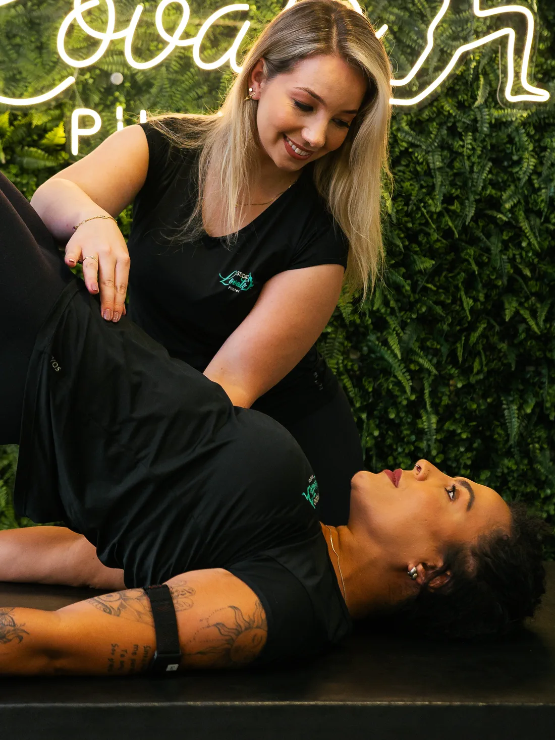 Pricila Lovato ajoelhada no chão do studio, com as mãos guiando uma aluna deitada de costas no tapete; ao fundo, o letreiro de neon do Studio Lovato Pilates.