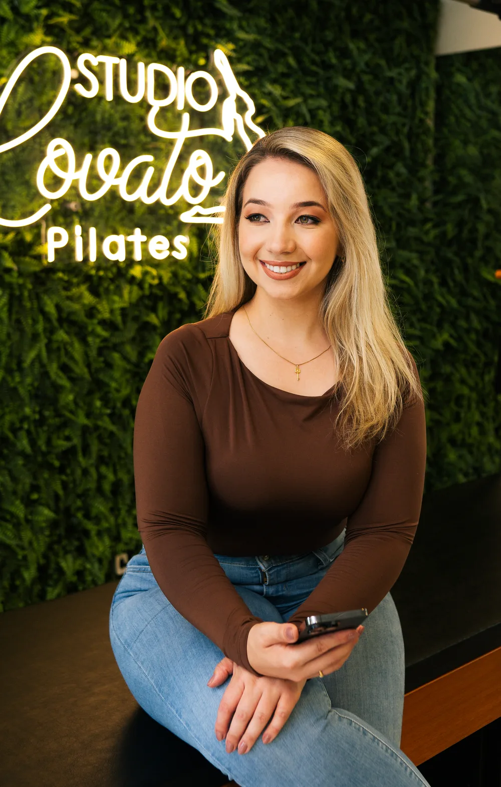 Pricila Lovato sentada no studio, de blusa marrom e jeans, com o celular na mão e o letreiro de neon do Studio Lovato Pilates ao fundo.